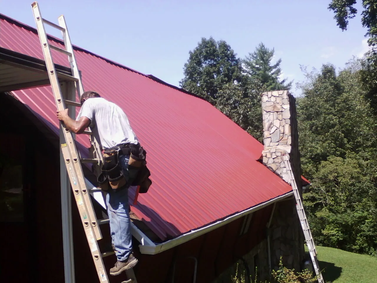 Roofer on a ladder beside a red metal roof and stone chimney — Soffit & Fascia Repair by Roofing Brothers in Paris